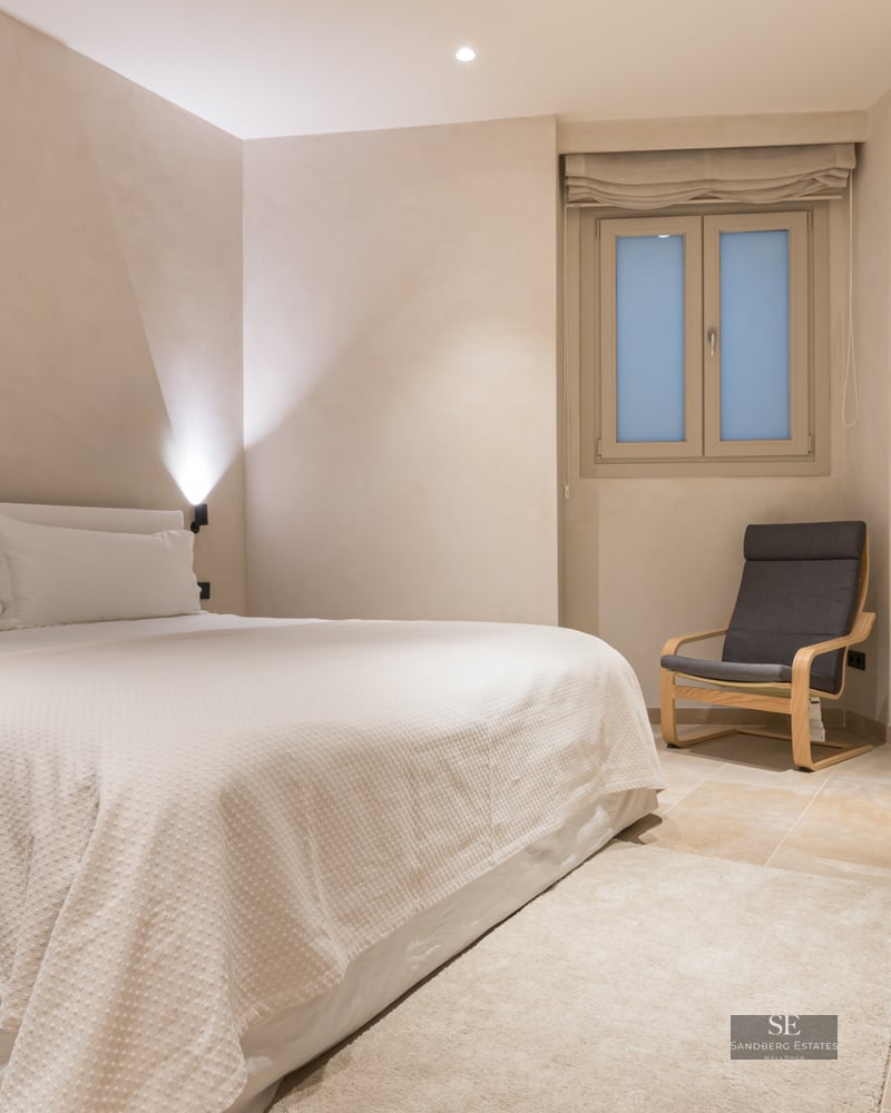 A serene bedroom with a large white bed, a grey designer chair, stone flooring, and soft beige walls.