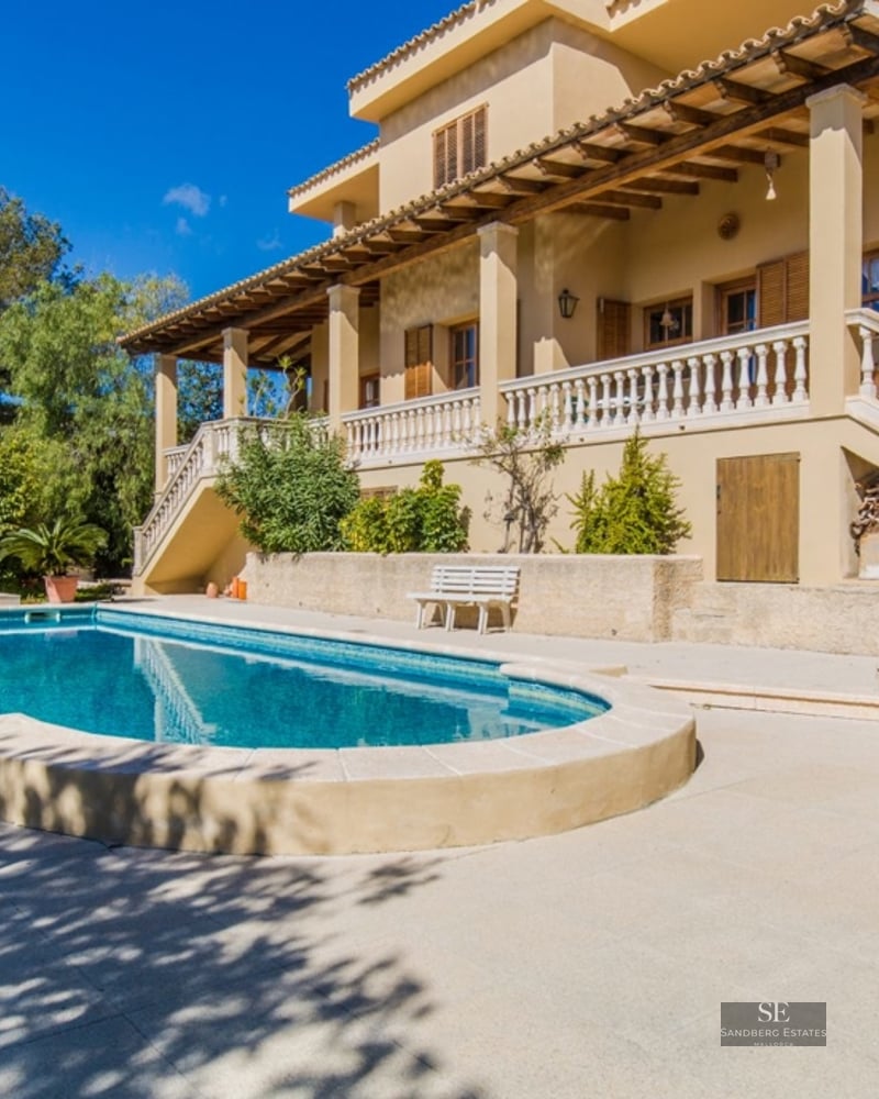 Large swimming pool in front of a multi-story Mediterranean villa with a terrace and lush green hills under a blue sky.