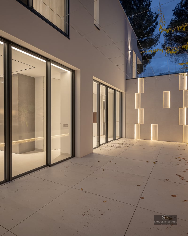 Modern villa terrace at night featuring large glass sliding doors, integrated LED wall lighting, and light stone tiling.
