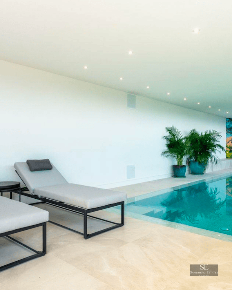 Modern indoor swimming pool with turquoise water, grey sun loungers, and a large scenic coastal wall mural.