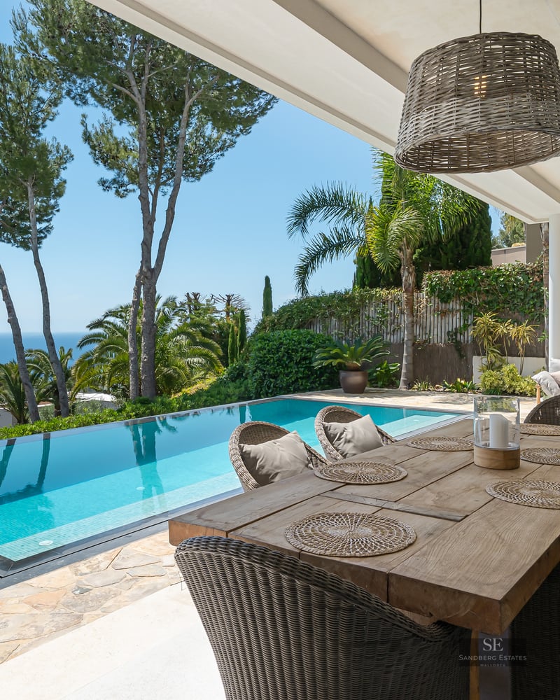 A covered terrace with a wooden dining table next to an infinity pool overlooking the blue sea and pine trees.