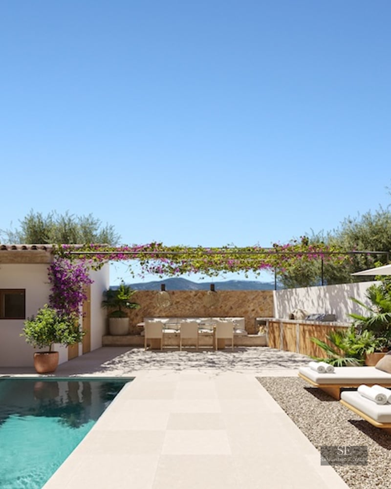 Modern turquoise pool with wooden sun loungers, outdoor dining under a flowering pergola, and rustic stone walls.
