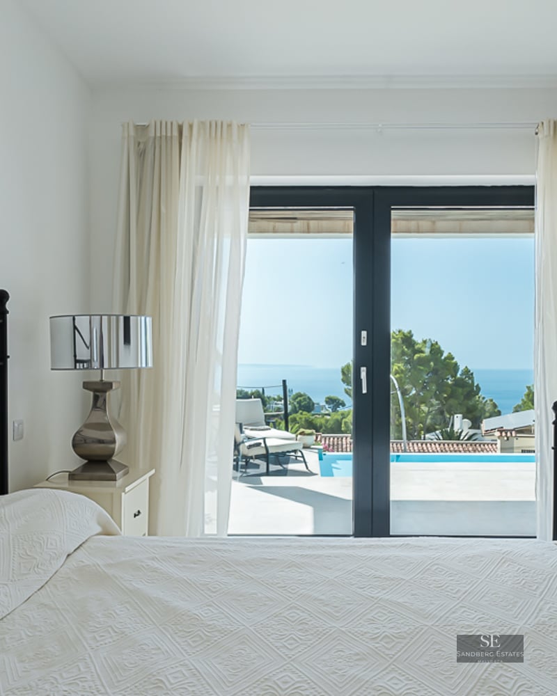 White bedroom with a black wrought iron bed and glass doors opening to a sunny terrace with a pool and sea view.