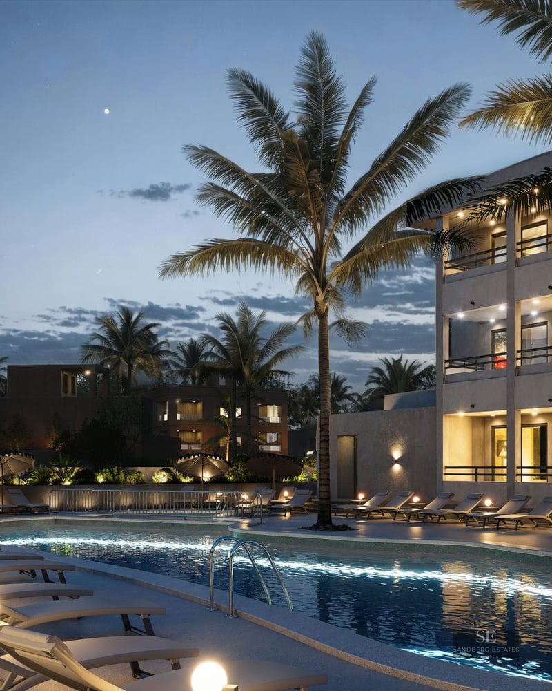 Illuminated swimming pool surrounded by sun loungers and palm trees under a twilight sky next to a modern building.