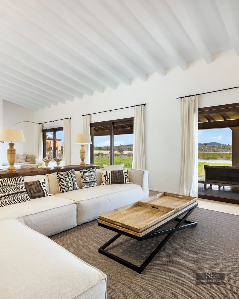 Bright living room with large L-shaped sofa, white wooden ceiling beams, and glass doors opening to a scenic terrace.