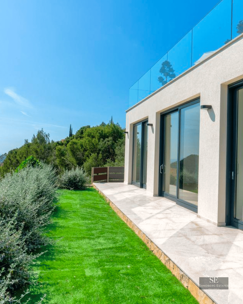 Modern white villa with large glass sliding doors, stone patio, green lawn, and panoramic sea and mountain views.