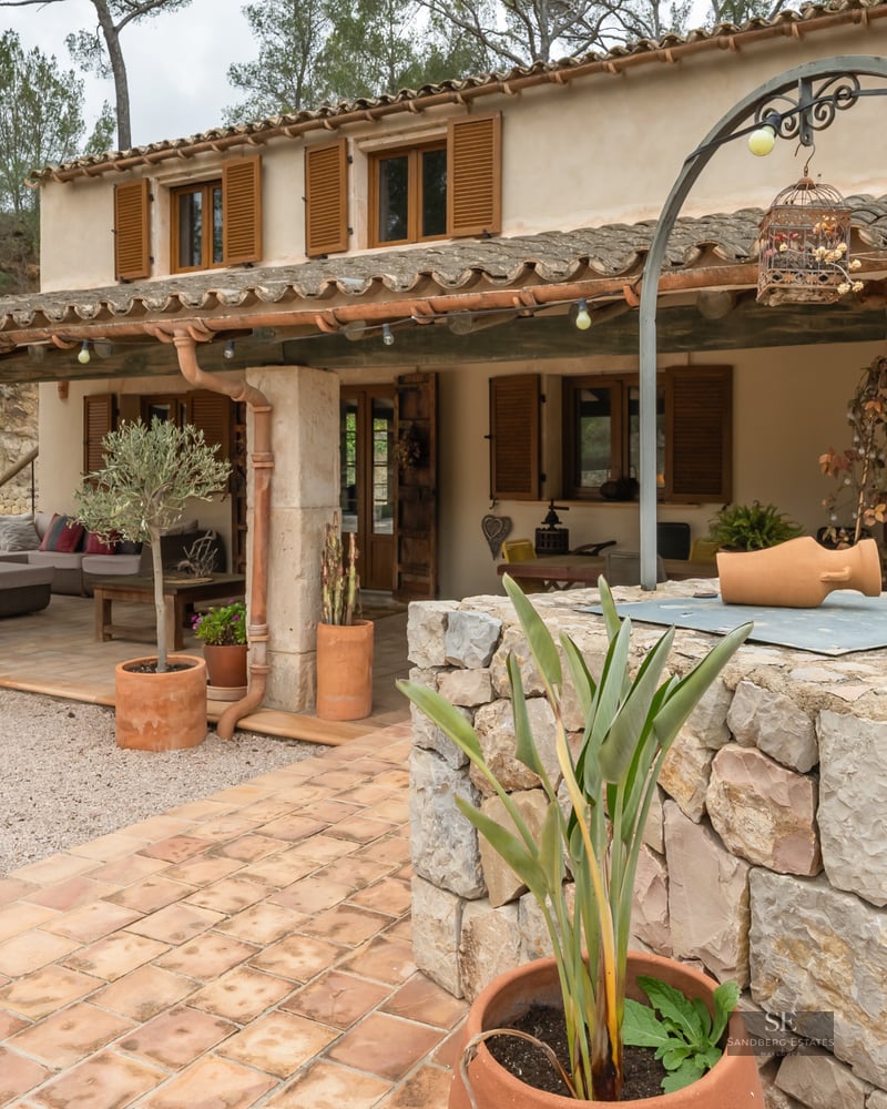Facade of a rustic house with a covered porch, wooden shutters, and a stone well in the foreground.
