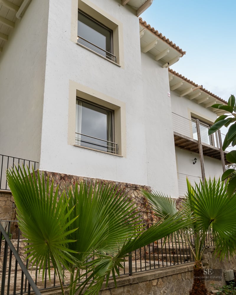 Three-story white villa facade with stone foundation, exterior stairs, and palm trees under a clear sky.