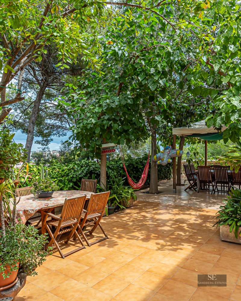 Large tiled terrace surrounded by mature trees featuring two wooden dining areas and a red hammock in the shade.