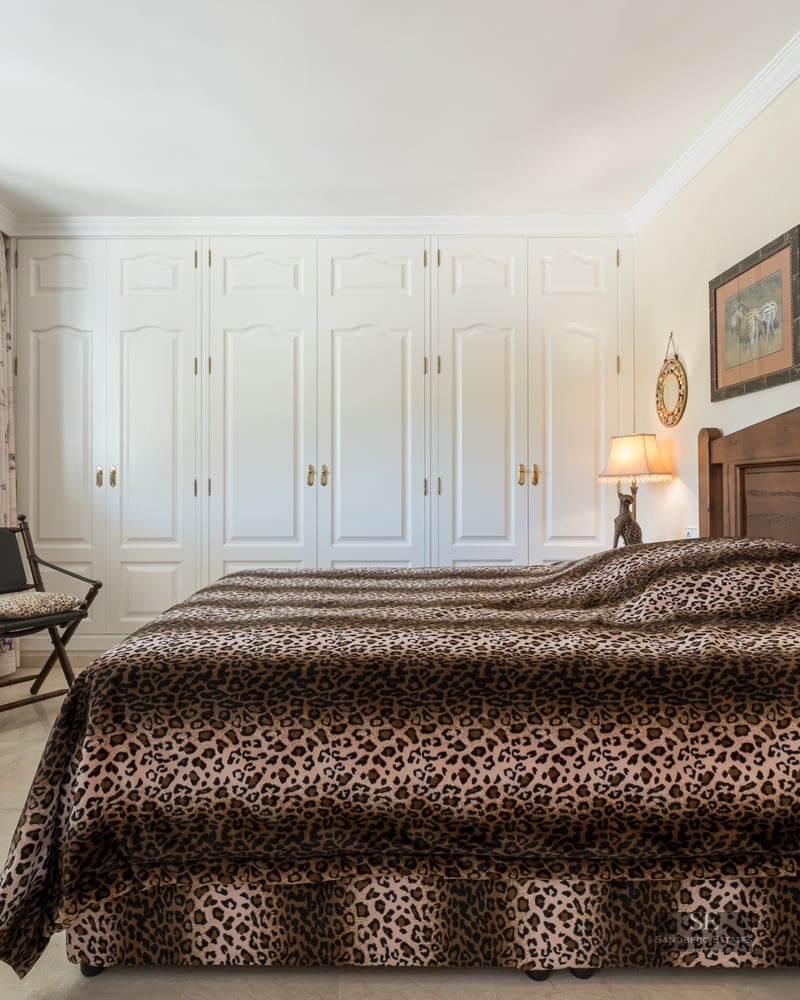 Bedroom featuring leopard print bedding, dark wood furniture, and a full wall of white built-in wardrobes.