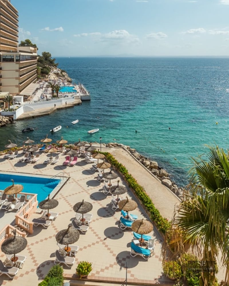 Aerial view of a swimming pool area with sunbeds and umbrellas overlooking a clear turquoise bay and rocky coastline.