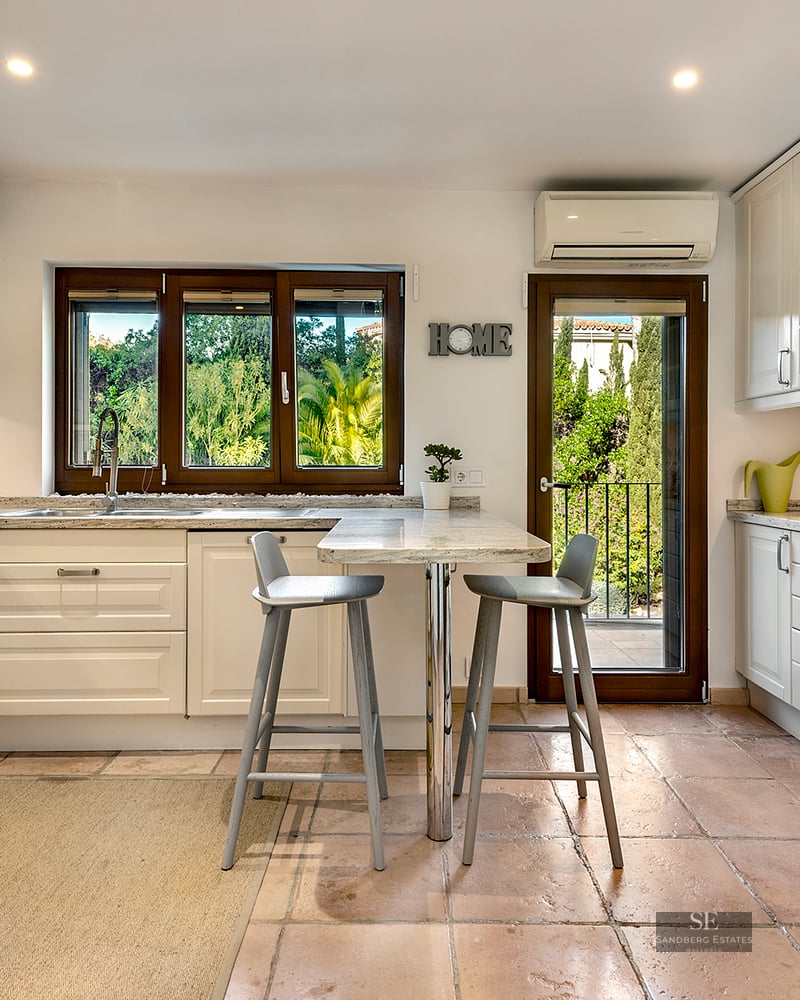 Bright kitchen with cream cabinets, granite counters, terracotta floors, breakfast bar, and window view of lush greenery.