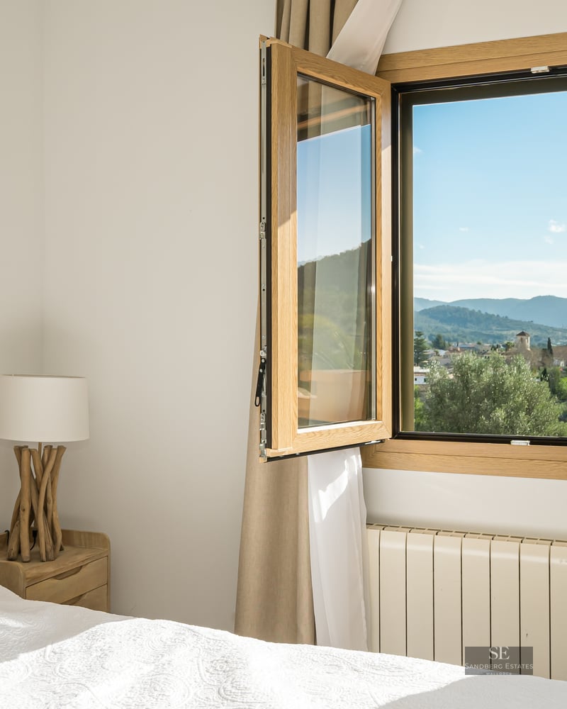 Cozy bedroom with tufted headboard and open window showing panoramic views of a village and mountains.