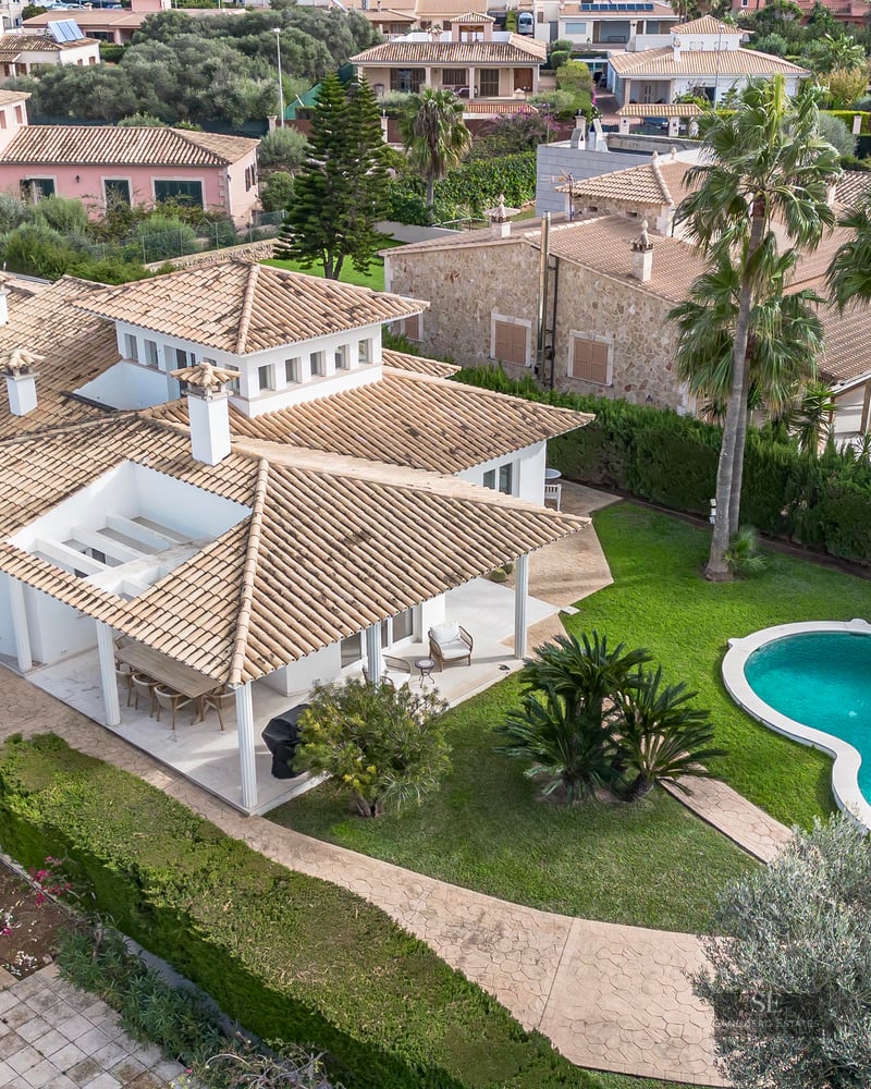 Aerial view of a white villa with a terracotta roof, a turquoise swimming pool, and a lush green garden.