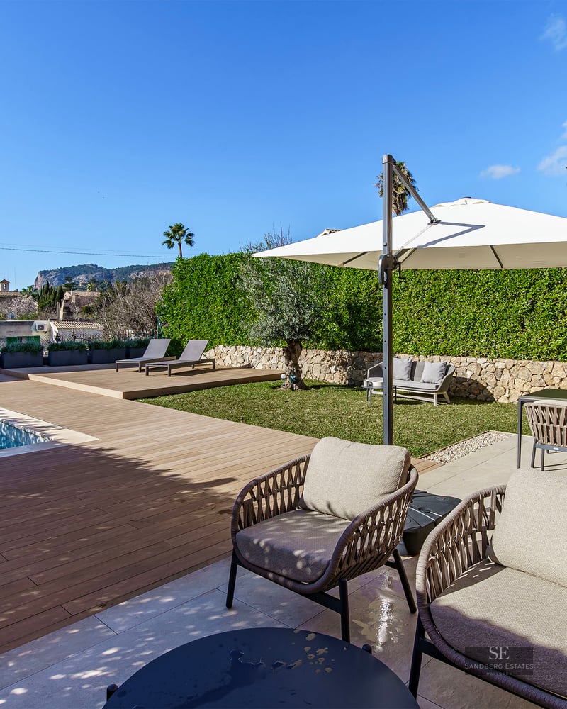 Modern swimming pool with wooden deck, stylish outdoor furniture, and lush green hedges under a clear blue sky.
