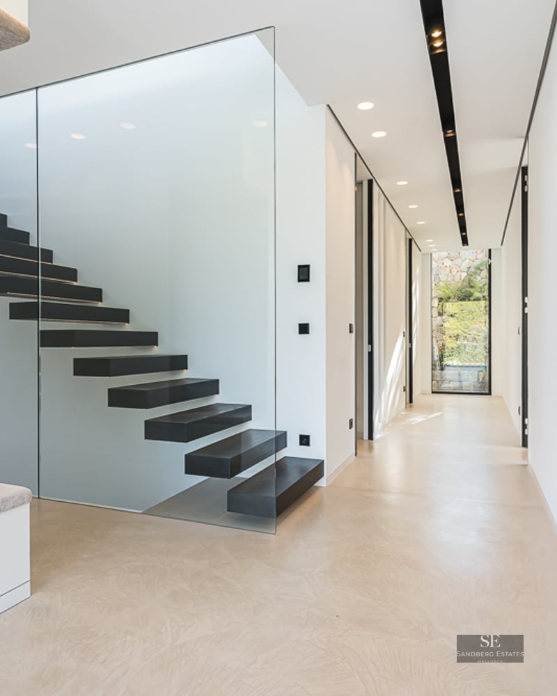 Modern minimalist hallway featuring a black floating staircase behind a glass panel and sleek recessed lighting.