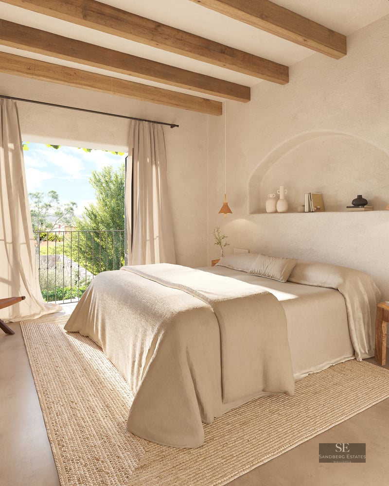 Bright bedroom featuring a large bed, wooden ceiling beams, an arched wall niche, and a balcony viewing lush trees.
