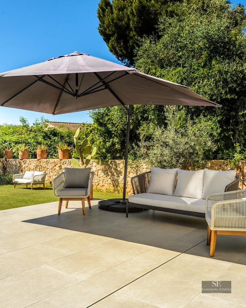 Sun-drenched terrace with modern grey sofa, armchairs, and large parasol next to a pool and lush garden.