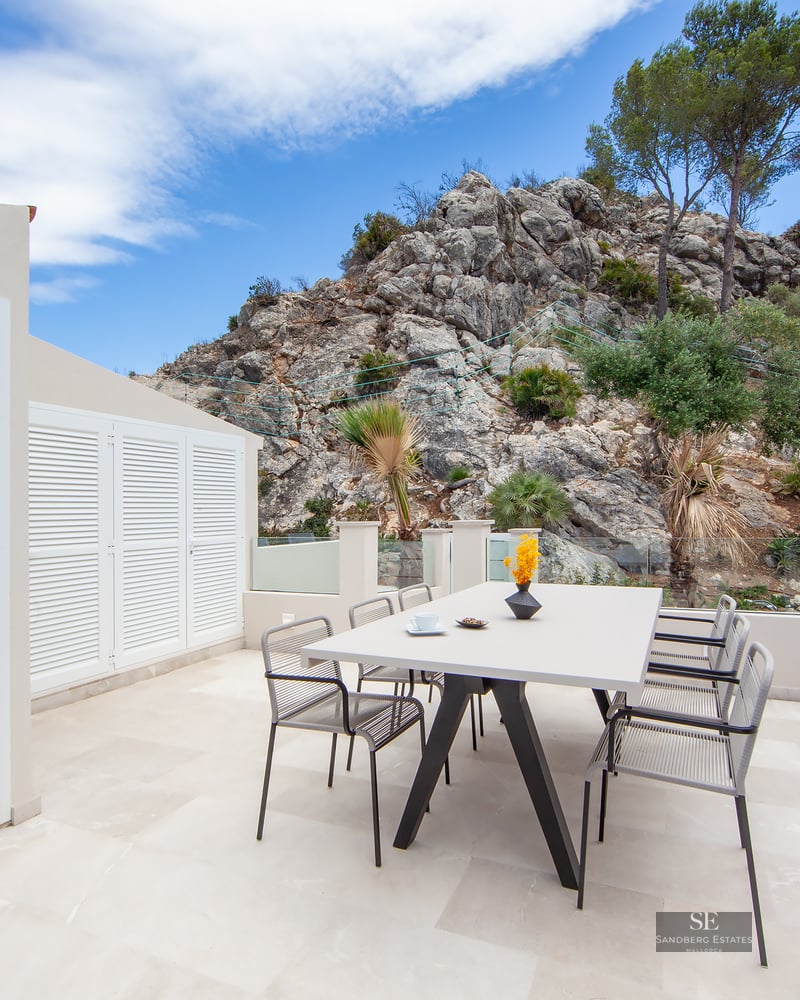 A modern white terrace with a dining table, chairs, and a barbecue area, set against a rocky mountain backdrop.