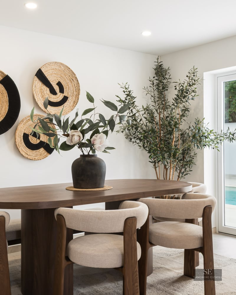 Oval wooden dining table with beige chairs in front of glass doors showing a swimming pool.