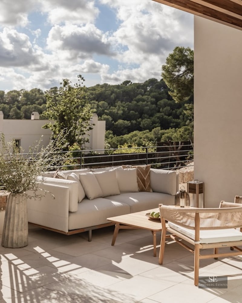 Outdoor terrace with modern sofa, wooden table, and woven chairs overlooking a lush green valley.