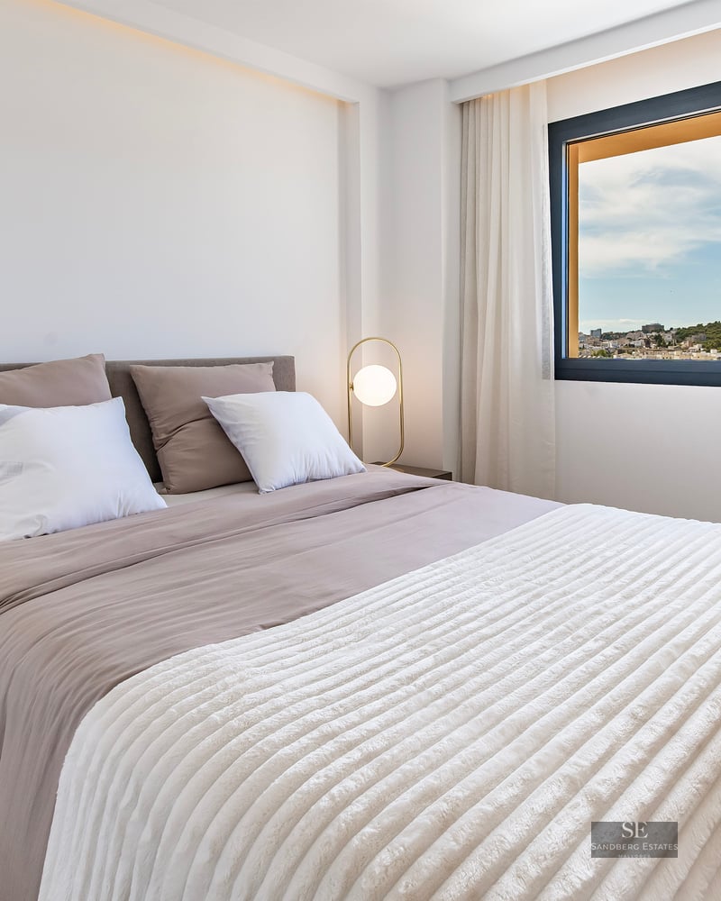 Modern bedroom with a large bed, gold designer lamps, and a window overlooking a city landscape.