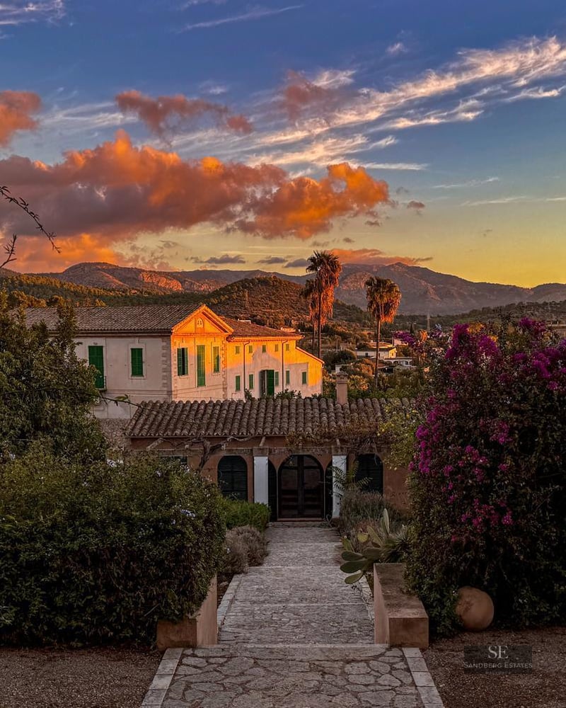 Scenic view of a white Mediterranean villa with terracotta roof at sunset, surrounded by lush gardens and mountains.