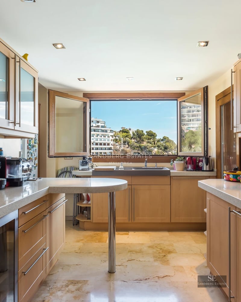 Modern kitchen with light wood cabinets, marble floors, and a large window overlooking a coastal landscape.