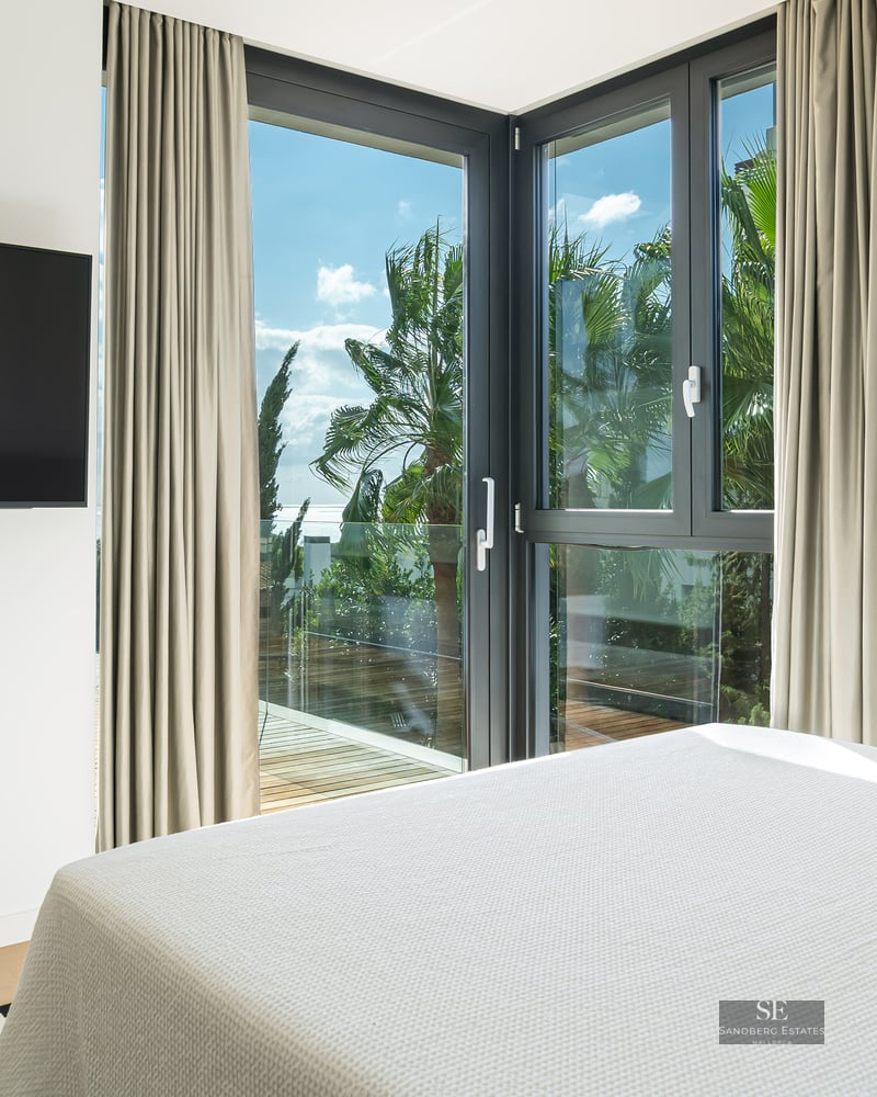Bright bedroom with a large bed, wall-mounted TV, and corner windows looking out to palm trees and a sunny sky.
