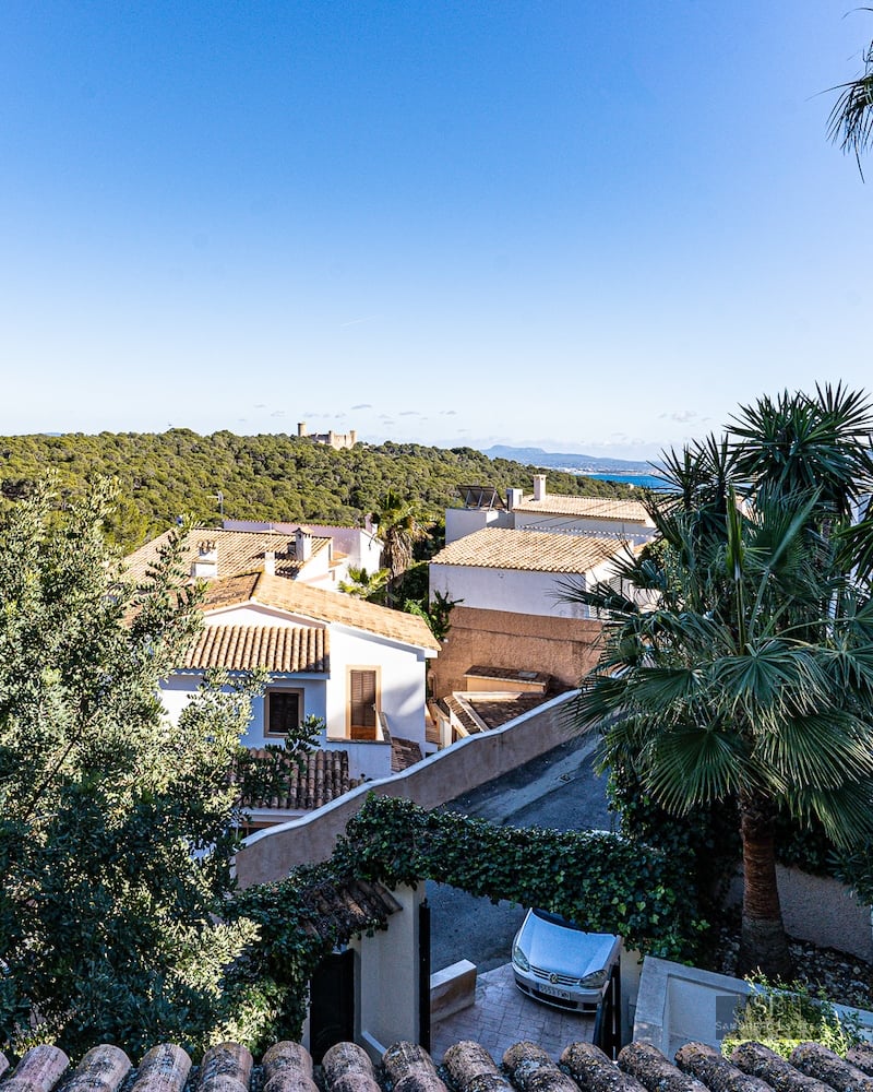 Elevated panoramic view showing white villas, lush greenery, a distant castle on a hill, and the sea under a blue sky.