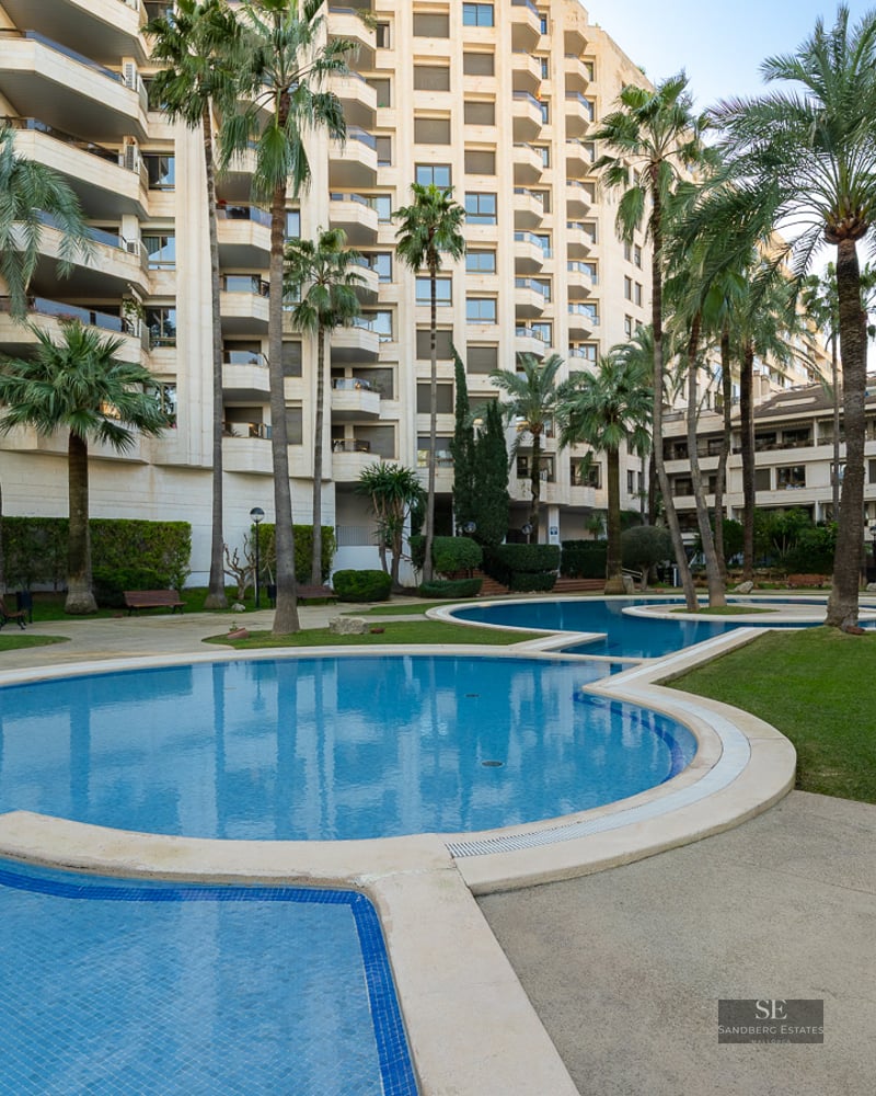 Winding blue swimming pool surrounded by green grass and tall palm trees in front of a modern apartment building.