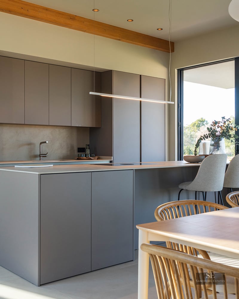 Minimalist kitchen with taupe island, integrated appliances, and wooden dining table under exposed beams.