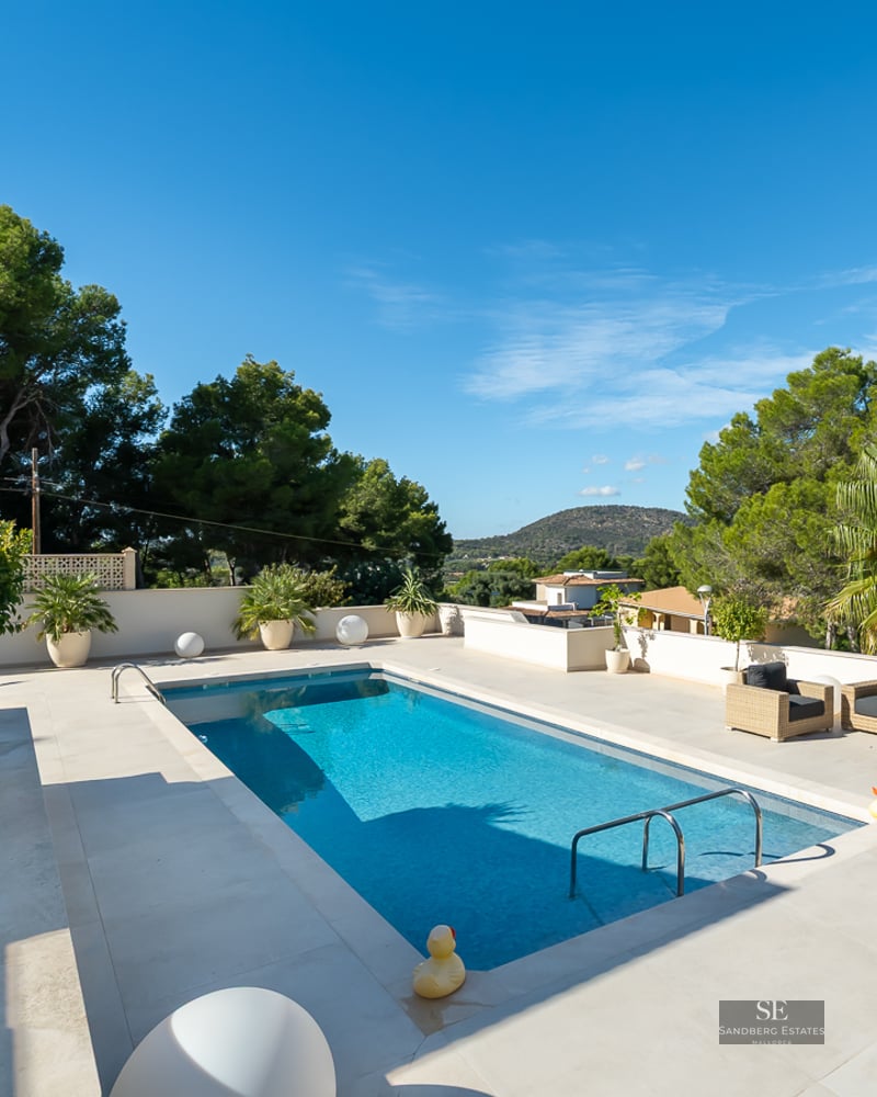 Piscine bleue rectangulaire sur terrasse blanche avec mobilier de jardin et vue sur la forêt méditerranéenne.