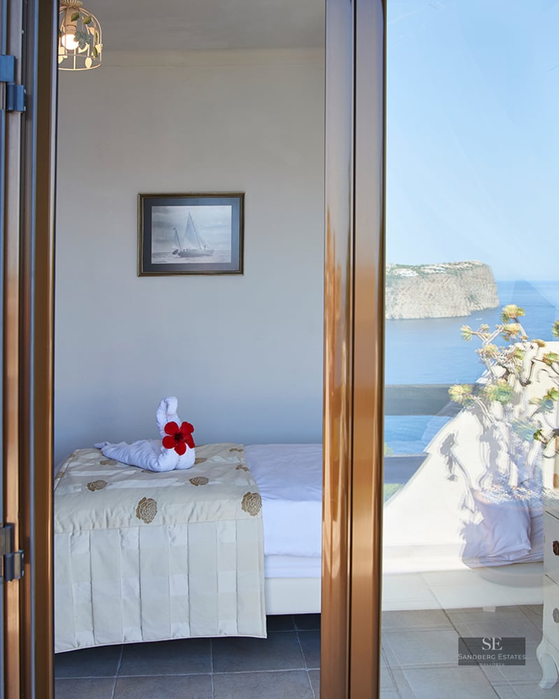 View through shutters of a bedroom with a single bed and towel swan, looking out to the blue sea and cliffs.
