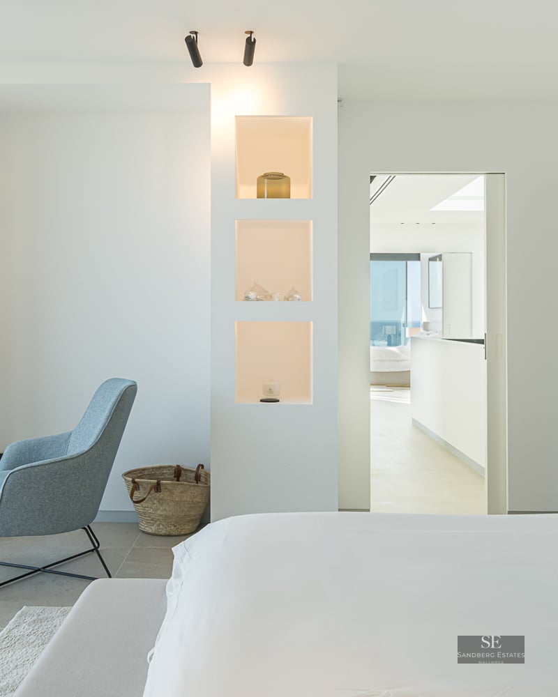Bright modern master bedroom featuring a grey bed, blue designer armchair, and built-in illuminated wall shelving.