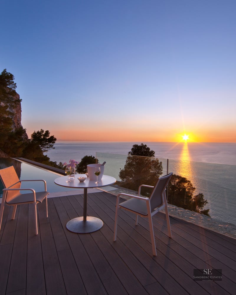 Sunset view from a luxury terrace with an infinity pool, wooden decking, and white modern outdoor seating.