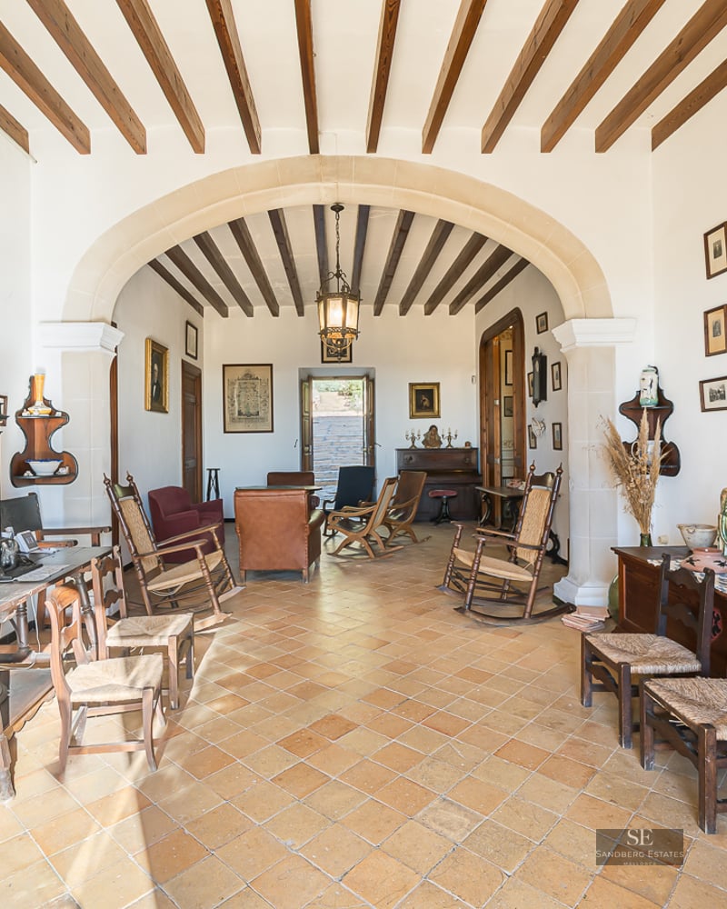 Spacious living room with terracotta floors, exposed wooden beams, a large stone archway, and rustic antique furniture.