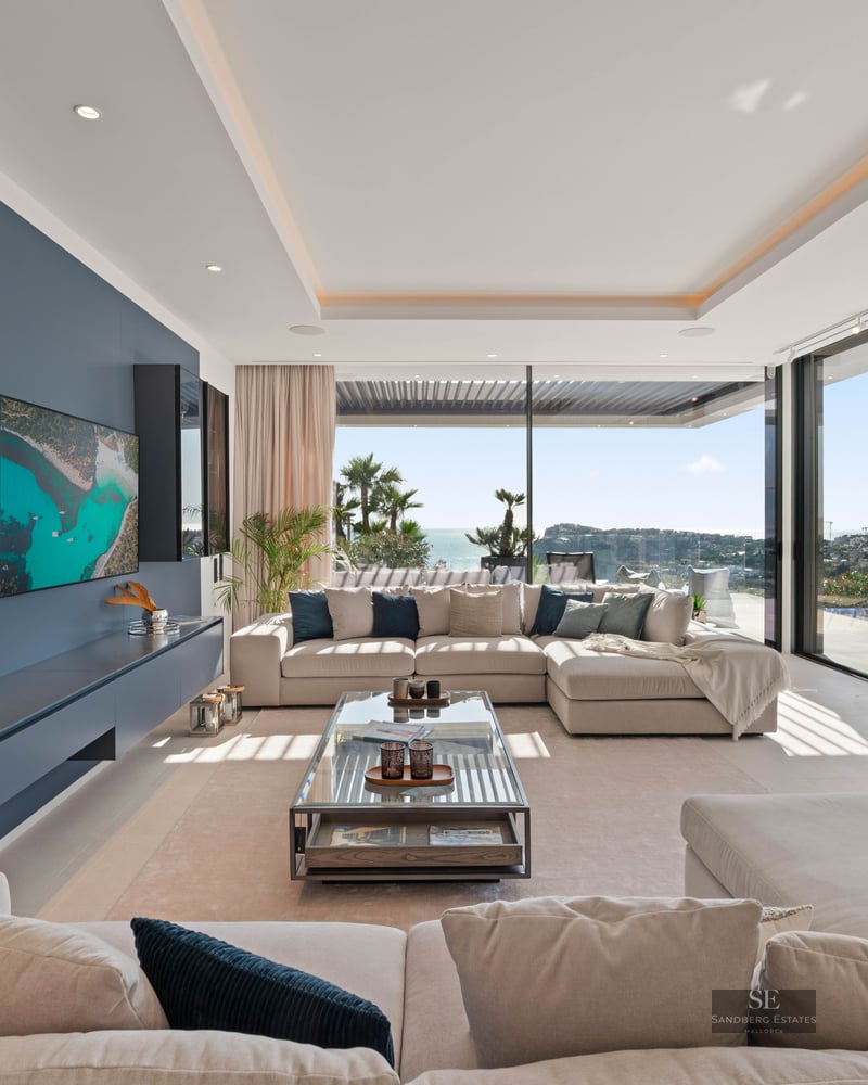 Spacious modern living room with beige sofas, a blue accent wall, and large windows overlooking the sea and an infinity pool.