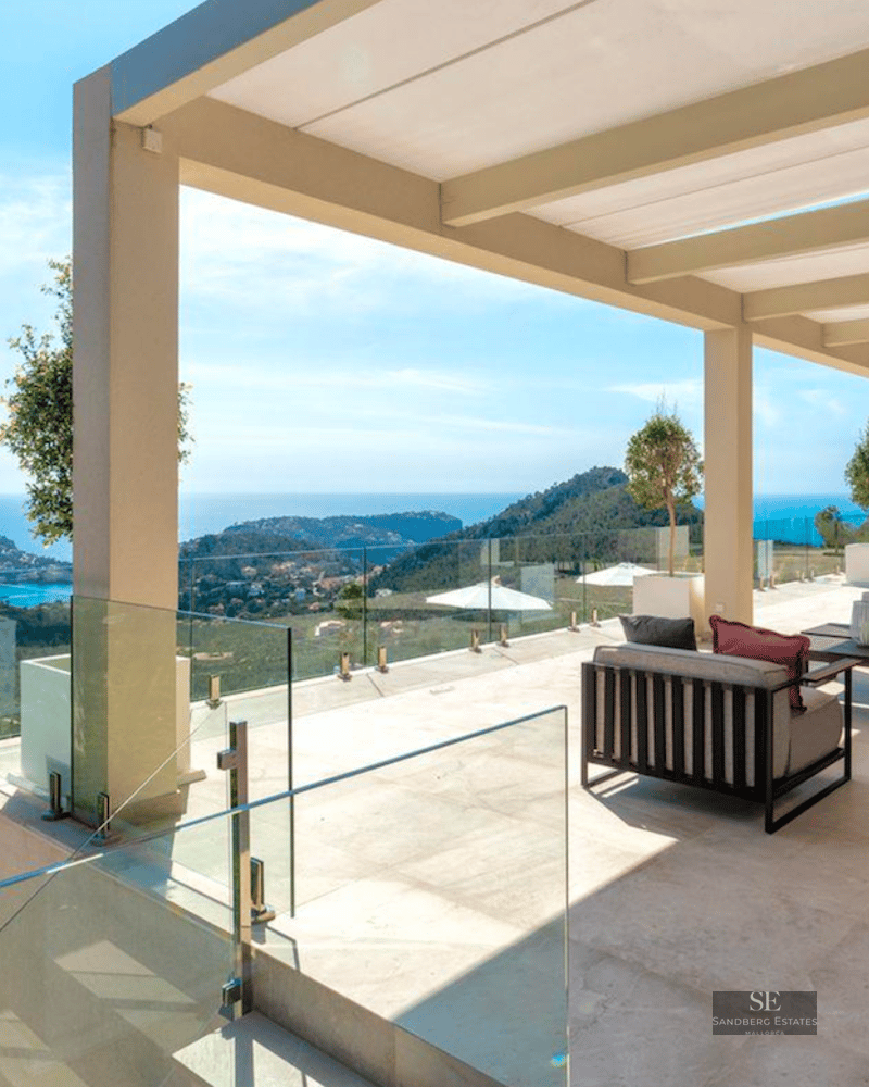 Large modern terrace with grey sofas, glass railings, and a stunning view of a blue bay and green hills.