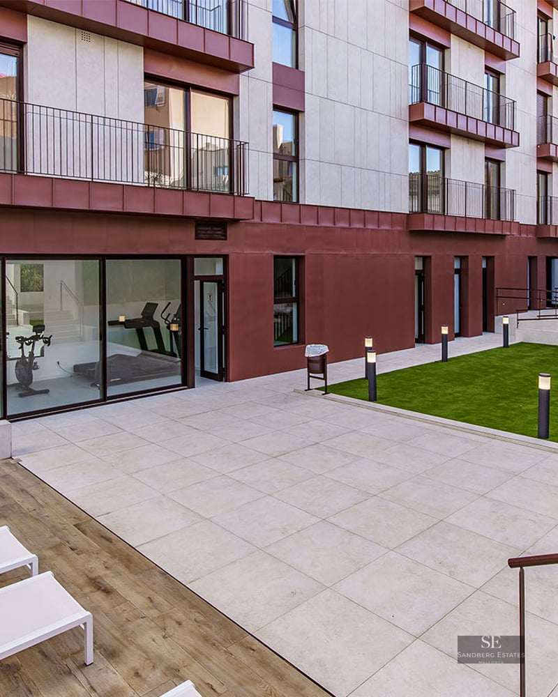 Courtyard with stone tiles, sun loungers, a lawn area with sofas, and a glass-walled gym at a modern building.