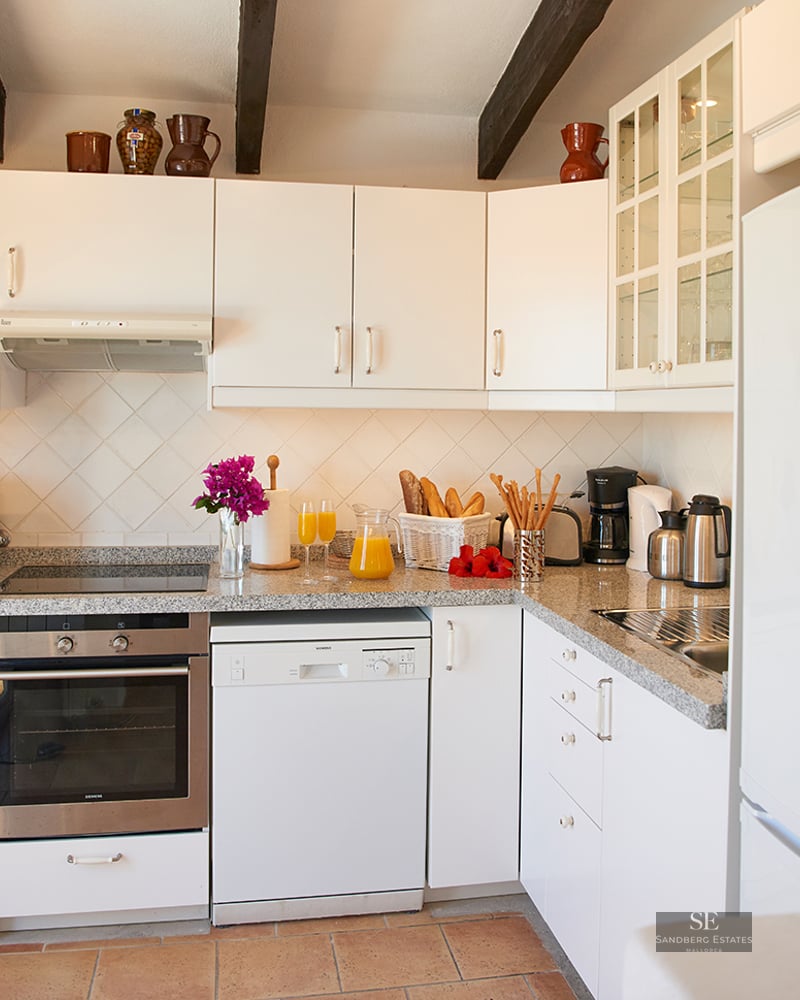 Bright kitchen with white cabinets, dark wood beams, terracotta floor, and modern appliances.