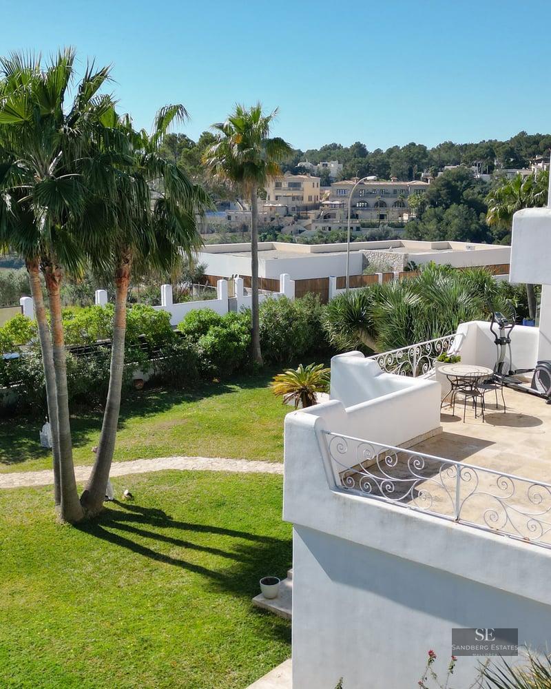 High-angle view of a white villa with a terrace, garden with palm trees, and views of the blue sea and cliffs in the distance.