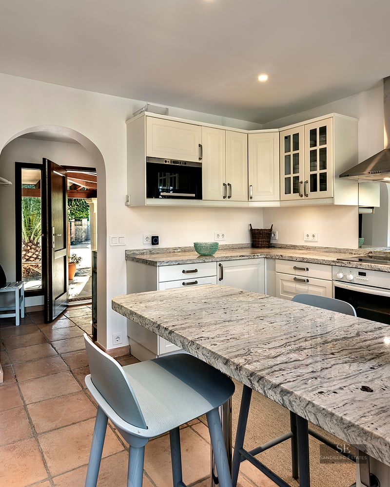 Bright kitchen with white cabinets, granite countertops, terracotta floors, and an archway leading to a sunny exterior.