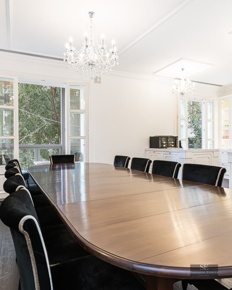 Large wooden dining table with black velvet chairs under crystal chandeliers in a bright, white room with a fireplace.