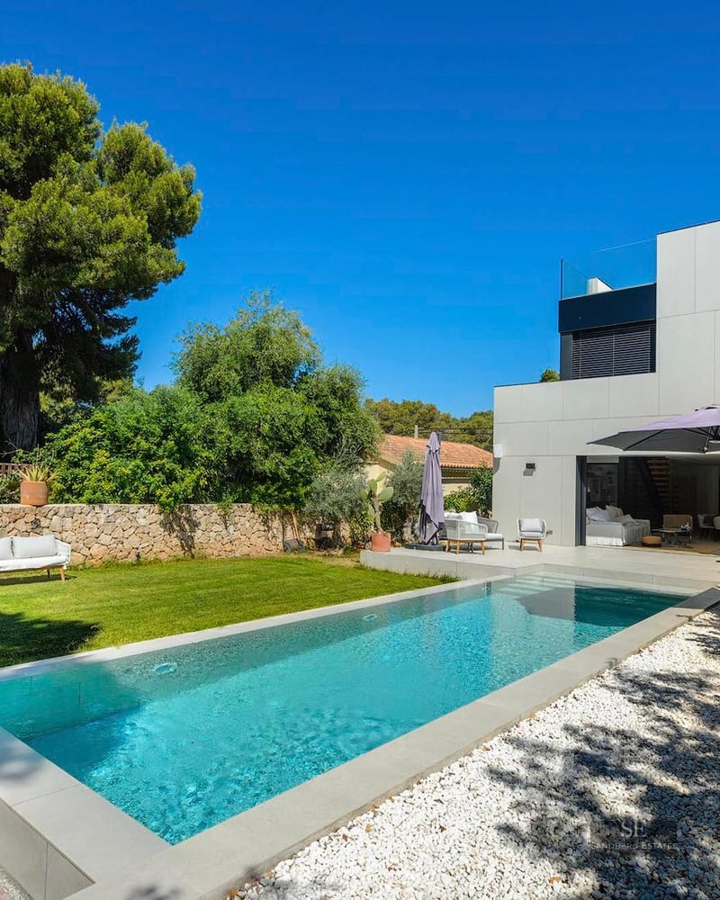 Rectangular swimming pool surrounded by a green lawn, white gravel, and a modern white villa under a clear blue sky.