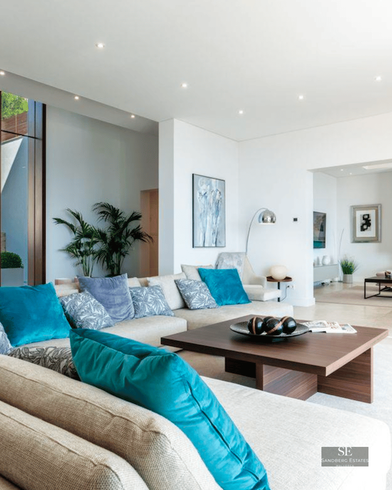 Spacious modern living room with beige sectional sofa, teal pillows, wooden table and floor-to-ceiling windows.