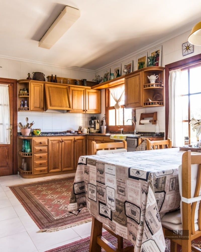 Bright rustic kitchen with wooden cabinets, a dining table with patterned cloth, and sunlight streaming through windows.