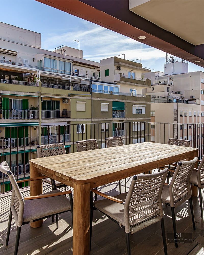 Sunlit terrace with wooden dining table, woven chairs, and black spiral staircase against a city backdrop.