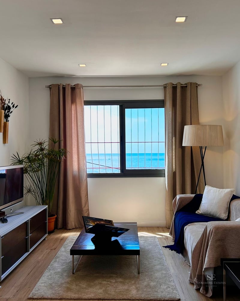 Modern living room featuring a beige sofa, wooden coffee table, and a large window overlooking the blue ocean.