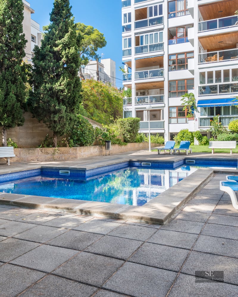 A rectangular blue swimming pool surrounded by stone tiles, sun loungers, and tall green trees next to an apartment block.